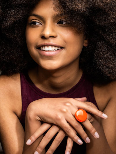 jeune femme peau foncée qui sourit, mains croisées avec bague pastille de verre orange