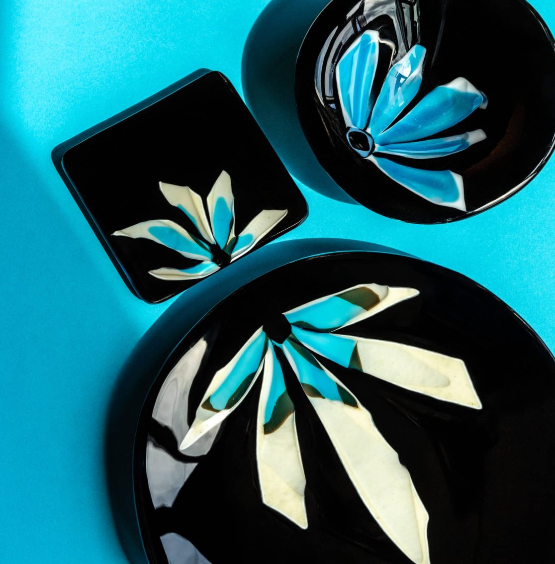 ensemble d'assiettes de verre noir avec pétales illustrant des demi fleurs aux couleurs lumineuses de blanc, crème et turquoise.